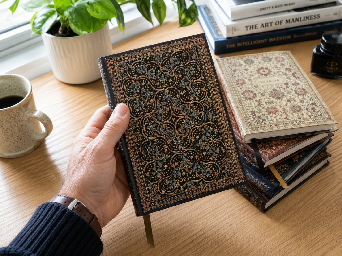 Ornate Paperblanks journal held above a wooden desk with coffee, books, and plants
